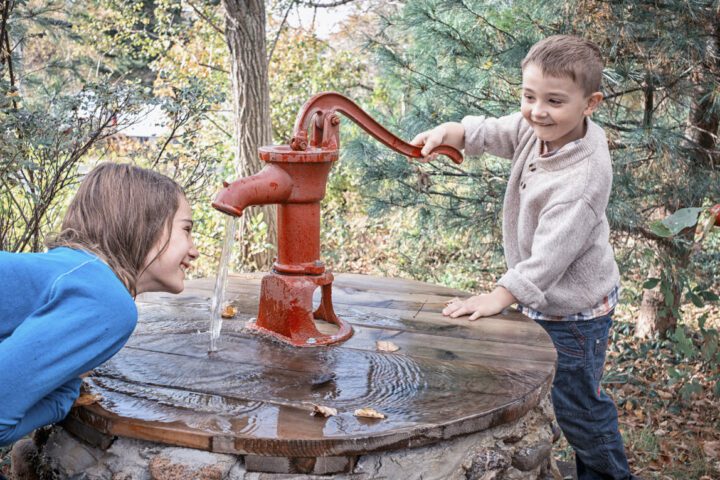 A young boy is having fun giving his sister a drink at an antique hand operated water pump. Forest background with late day or evening light. Well is stone, pump is red. It is early autumn. Children are smiling and laughing. Leica camera photograph.